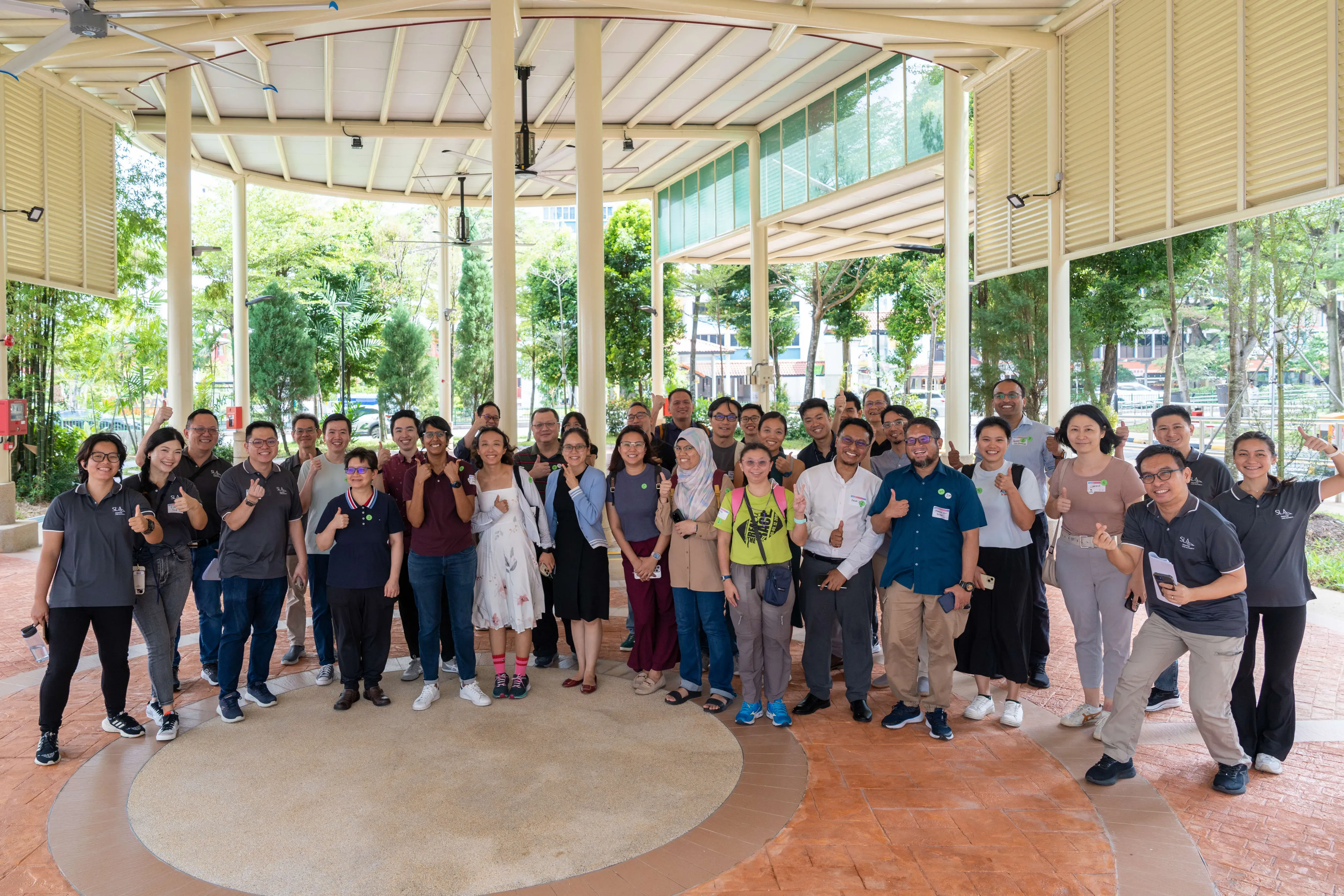 A large group of about 30 people posing and giving thumbs up under an open outdoor pavilion surrounded by trees.